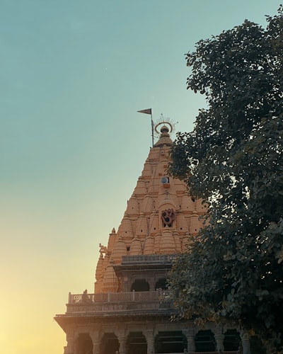 A majestic temple spire with intricate carvings rises into the clear sky, with a sacred symbol adorning its facade. A single flag atop the spire adds a sense of reverence. The warm sunlight bathes the temple in a gentle glow, contrasting with the deep green of the surrounding tree foliage.