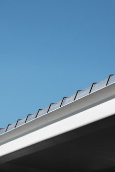 A minimalistic view of a bridge or building edge against a clear blue sky. The structure features a row of transparent panels or railings, creating a geometric and modern look.