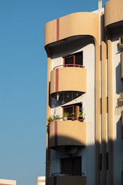 A modern apartment building with a distinct architectural style is visible. The structure features rounded balconies adorned with potted plants and hanging decorations, with a warm sunlight casting shadows on the facade. The building is painted in earth tones and red accents, and it stands against a clear blue sky.