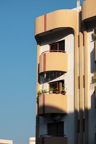 A modern apartment building with a distinct architectural style is visible. The structure features rounded balconies adorned with potted plants and hanging decorations, with a warm sunlight casting shadows on the facade. The building is painted in earth tones and red accents, and it stands against a clear blue sky.