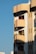 A modern apartment building with a distinct architectural style is visible. The structure features rounded balconies adorned with potted plants and hanging decorations, with a warm sunlight casting shadows on the facade. The building is painted in earth tones and red accents, and it stands against a clear blue sky.