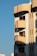 A modern apartment building with a distinct architectural style is visible. The structure features rounded balconies adorned with potted plants and hanging decorations, with a warm sunlight casting shadows on the facade. The building is painted in earth tones and red accents, and it stands against a clear blue sky.