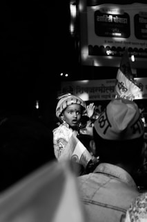 A child wearing traditional attire and a turban is amidst a crowd, holding a small Indian flag. The image is in black and white, creating a nostalgic ambiance. The child looks curious or surprised, while the crowd seems focused on an event. There are visible signs and banners in the background.