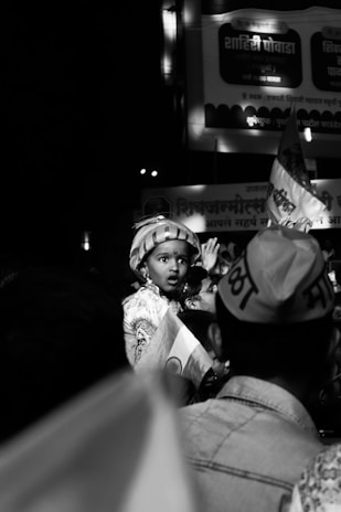 A child wearing traditional attire and a turban is amidst a crowd, holding a small Indian flag. The image is in black and white, creating a nostalgic ambiance. The child looks curious or surprised, while the crowd seems focused on an event. There are visible signs and banners in the background.