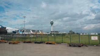 An airport tarmac with a parked airplane on the left side, surrounded by a fence with a control tower visible in the background. The sky is mostly cloudy with patches of blue. Luggage carts are lined up near the fence, and the area seems secure with barbed wire on the fence.