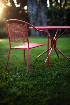 A metal chair with a mesh back and seat is positioned beside a matching round table. The furniture is set on lush green grass. Sunlight filters through trees in the background, creating a warm glow and soft bokeh effect.