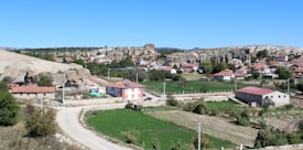 A rural landscape with a mix of small houses and agricultural fields, surrounded by rocky outcrops. The sky is clear and blue, providing a backdrop to the scene. Vegetation is scattered around the area, with patches of greenery and trees interspersed between the buildings.