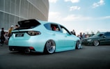 A mint green customized car with sleek, lowered suspension and shiny chrome wheels is parked on a street. It is surrounded by people and another car, creating a car meet atmosphere near a modern building on a clear day.