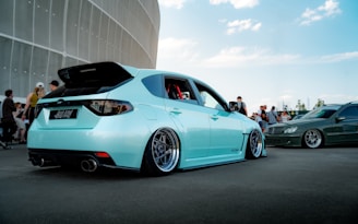 A mint green customized car with sleek, lowered suspension and shiny chrome wheels is parked on a street. It is surrounded by people and another car, creating a car meet atmosphere near a modern building on a clear day.