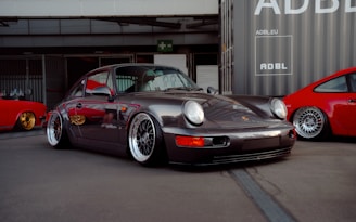 A sleek, classic sports car is prominently displayed, featuring a dark metallic finish and polished alloy wheels. The car is parked in an urban setting next to vibrant red sports cars. The scene is set against a backdrop of industrial buildings and shipping containers.