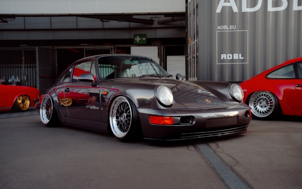 A sleek, classic sports car is prominently displayed, featuring a dark metallic finish and polished alloy wheels. The car is parked in an urban setting next to vibrant red sports cars. The scene is set against a backdrop of industrial buildings and shipping containers.