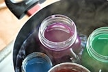 Several jars filled with different colored liquids are submerged in a pot of boiling water. The pot handle is visible at the edge, and steam rises from the water.