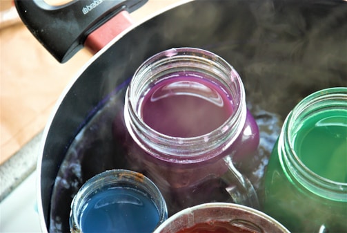 Several jars filled with different colored liquids are submerged in a pot of boiling water. The pot handle is visible at the edge, and steam rises from the water.