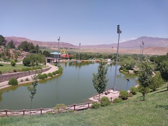 A serene park landscape featuring a large pond surrounded by lush green grass and trees. Several tall lighting poles border the pond, and a small promenade provides a walking path. In the distance, a colorful playground structure is visible, and beyond that, mountains stretch across the horizon under a clear blue sky.