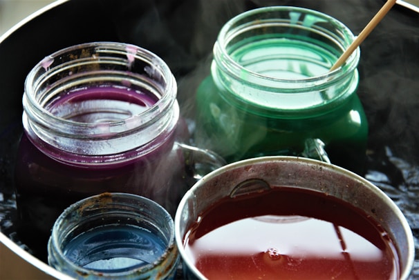 Four glass jars are filled with different colored liquids: purple, green, blue, and red. Each jar is open, releasing steam, and a wooden stick is dipping into the green liquid.