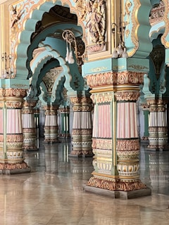 Intricate architectural columns and arches feature detailed carvings and pastel colors, creating an ornate and historical atmosphere. The design elements include floral and geometric patterns, with a polished floor reflecting the structural details.