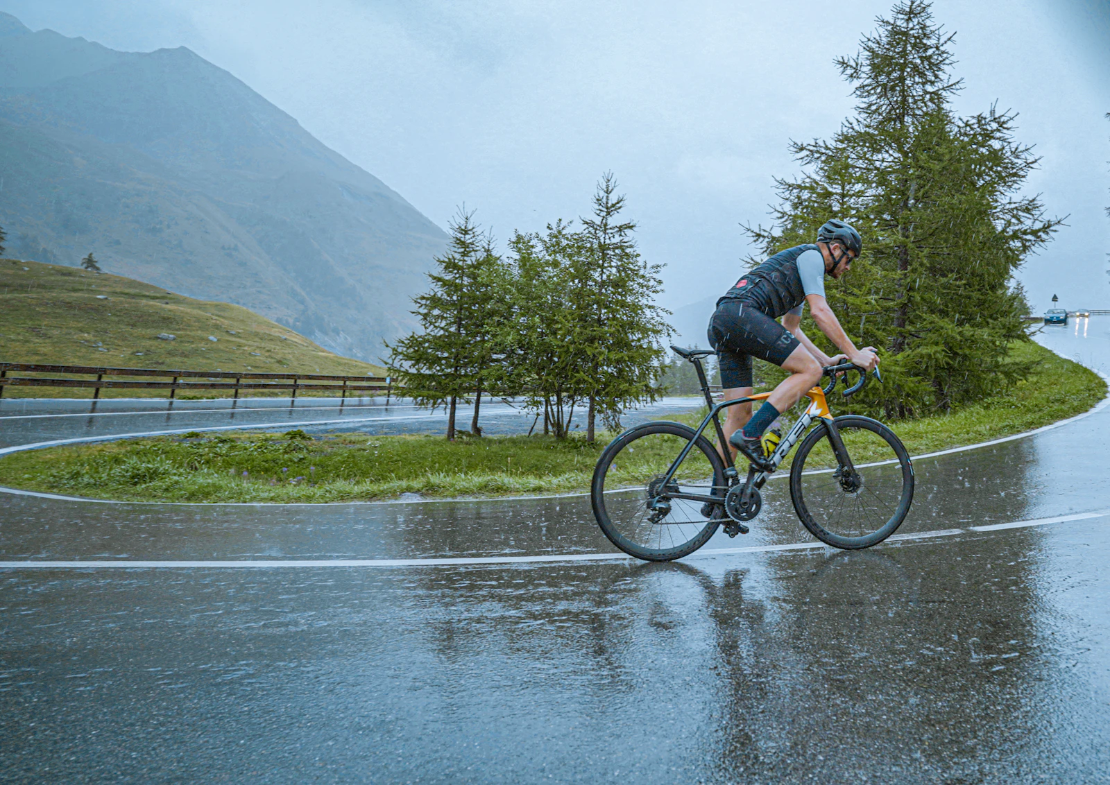A road cyclist climbing a wet mountain hairpin in the rain beneath pine trees