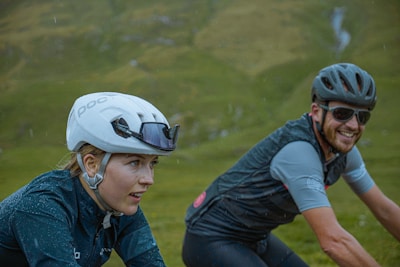 Two cyclists, wearing helmets and cycling gear, are riding in a lush, green outdoor setting. One cyclist appears focused while the other cyclist is smiling, suggesting enjoyment.