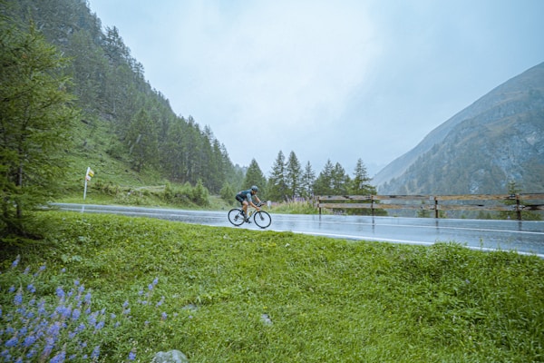 A cyclist rides along a wet road amidst a lush, mountainous landscape. The scene is surrounded by dense green trees and distant mist-covered mountains. The sky is overcast, suggesting recent or ongoing rainfall. The road is bordered by vibrant green grass and scattered purple flowers.