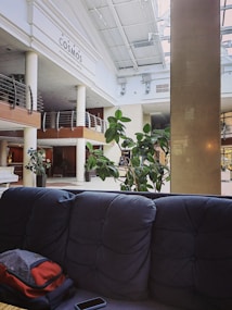 A modern hotel lobby area featuring a high ceiling with large windows allowing natural light to flood the space. There's a blue tufted couch in the foreground, and a backpack is placed on it. A potted plant adds greenery to the scene, and there are various architectural accents like wooden railings and a large pillar. The name 'Cosmos Petrozavodsk Hotel' is visible on the wall.