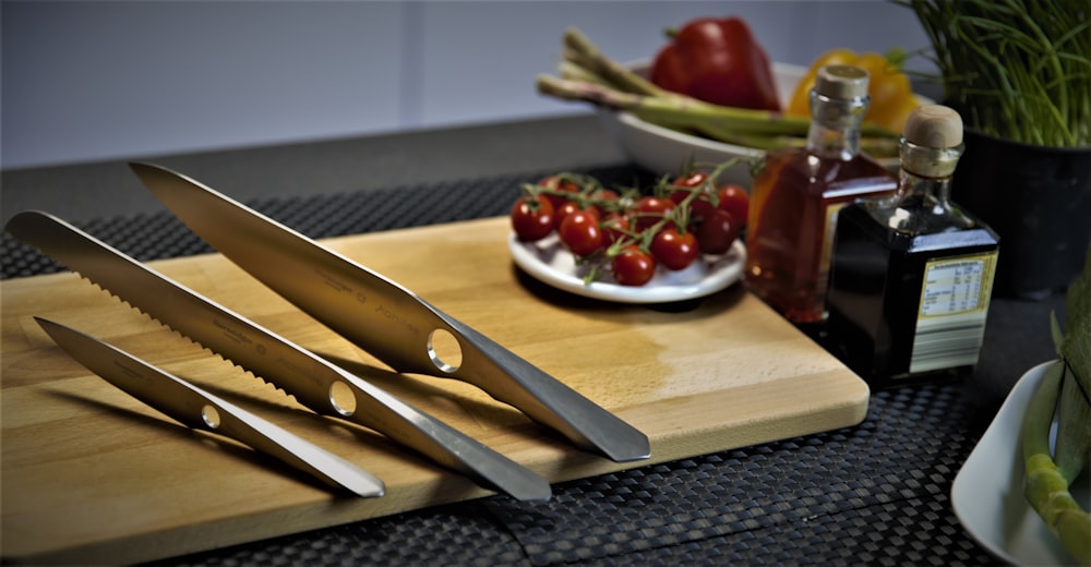 a cutting board topped with three knives next to a bowl of fruit