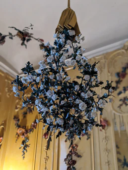 A decorative floral arrangement is hanging from the ceiling, featuring intricately designed white roses with green leaves set against a lavish, ornately decorated background. The background has golden hues with additional floral motifs, enhancing the luxurious ambiance.