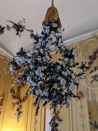 A decorative floral arrangement is hanging from the ceiling, featuring intricately designed white roses with green leaves set against a lavish, ornately decorated background. The background has golden hues with additional floral motifs, enhancing the luxurious ambiance.
