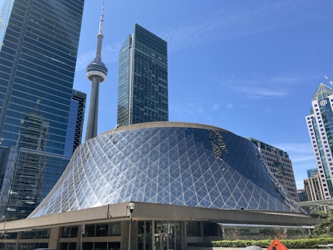 A modern architectural scene featuring a large, uniquely designed building with a glass and metal dome structure surrounded by tall skyscrapers. The iconic CN Tower is visible in the background against a clear blue sky, adding to the urban skyline.