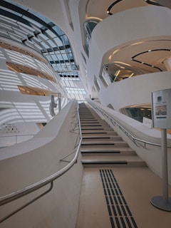 An interior view of a modern architectural space with sleek, curving lines. A wide staircase ascends between large, curved walls that are interspersed with expansive glass windows, allowing light to cascade through the space creating dynamic shadows. The design is futuristic and minimalistic, featuring metallic railings and a neutral color palette. A signpost with a conference announcement can be seen on one side.