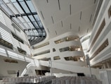 The image depicts a modern, spacious architectural interior with white and grey tones. Large, curved structures with multiple levels and glass elements are visible, creating an open, futuristic ambiance. Rows of empty chairs are aligned facing a stage or presentation area. Sunlight casts geometric shadows across the floor and walls, adding depth and complexity to the space.