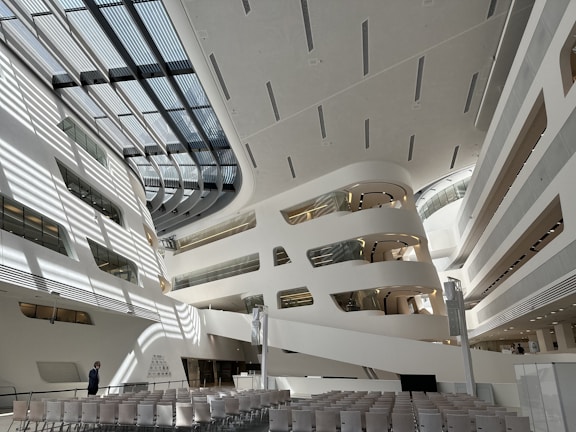 The image depicts a modern, spacious architectural interior with white and grey tones. Large, curved structures with multiple levels and glass elements are visible, creating an open, futuristic ambiance. Rows of empty chairs are aligned facing a stage or presentation area. Sunlight casts geometric shadows across the floor and walls, adding depth and complexity to the space.