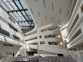 The image depicts a modern, spacious architectural interior with white and grey tones. Large, curved structures with multiple levels and glass elements are visible, creating an open, futuristic ambiance. Rows of empty chairs are aligned facing a stage or presentation area. Sunlight casts geometric shadows across the floor and walls, adding depth and complexity to the space.