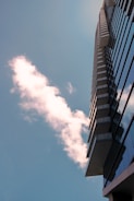 A modern high-rise building extends upward with reflective glass windows. A fluffy white cloud is seen against the backdrop of a clear blue sky.