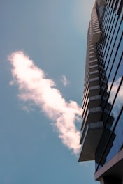A modern high-rise building extends upward with reflective glass windows. A fluffy white cloud is seen against the backdrop of a clear blue sky.