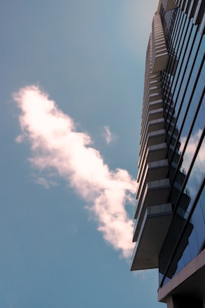 A modern high-rise building extends upward with reflective glass windows. A fluffy white cloud is seen against the backdrop of a clear blue sky.