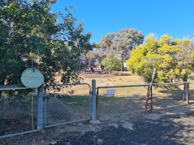 A rural scene with a chain-link fence in the foreground, featuring a 'private property' sign. Behind the fence, there is a yard with scattered trees and a playground structure with a slide. The sky is clear and blue, and the ground appears dry with patches of grass.