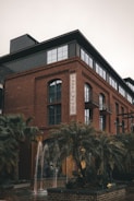 A multi-story brick building with large glass windows and a sign that reads 'THREE MUSES'. In the foreground, there are tall palm trees and a small fountain with water jets. The building has a modern industrial style with external metal fire escape stairs and balconies.
