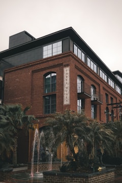 A multi-story brick building with large glass windows and a sign that reads 'THREE MUSES'. In the foreground, there are tall palm trees and a small fountain with water jets. The building has a modern industrial style with external metal fire escape stairs and balconies.