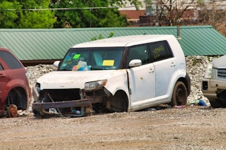 A white, heavily damaged car is parked on a gravel surface next to other vehicles. The car's front is missing parts, exposing components like the radiator. There are signs and notes stuck on the windshield, and the tires are absent. Surrounding the car are scattered debris and red and silver vehicles on either side.