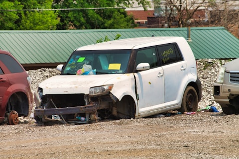 A white, heavily damaged car is parked on a gravel surface next to other vehicles. The car's front is missing parts, exposing components like the radiator. There are signs and notes stuck on the windshield, and the tires are absent. Surrounding the car are scattered debris and red and silver vehicles on either side.
