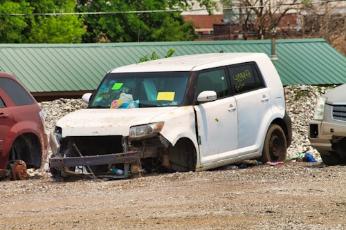 A white, heavily damaged car is parked on a gravel surface next to other vehicles. The car's front is missing parts, exposing components like the radiator. There are signs and notes stuck on the windshield, and the tires are absent. Surrounding the car are scattered debris and red and silver vehicles on either side.