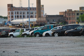 A row of abandoned or disused cars, parked on a gravel lot in an industrial area. The background features several old brick industrial buildings, one with a tall chimney. The cars appear worn and some have visible damage.