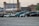 A row of abandoned or disused cars, parked on a gravel lot in an industrial area. The background features several old brick industrial buildings, one with a tall chimney. The cars appear worn and some have visible damage.