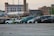A row of abandoned or disused cars, parked on a gravel lot in an industrial area. The background features several old brick industrial buildings, one with a tall chimney. The cars appear worn and some have visible damage.