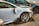 A damaged white car with significant front-end damage and a broken side mirror is parked among other wrecked vehicles. The tires and rims appear intact, while the front bumper and fender are severely dented, exposing internal components.