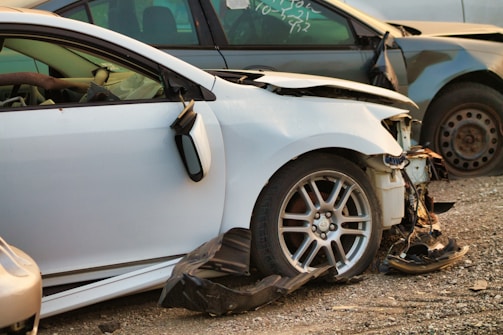 A damaged white car with significant front-end damage and a broken side mirror is parked among other wrecked vehicles. The tires and rims appear intact, while the front bumper and fender are severely dented, exposing internal components.