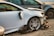 A damaged white car with significant front-end damage and a broken side mirror is parked among other wrecked vehicles. The tires and rims appear intact, while the front bumper and fender are severely dented, exposing internal components.