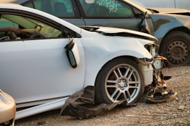 A damaged white car with significant front-end damage and a broken side mirror is parked among other wrecked vehicles. The tires and rims appear intact, while the front bumper and fender are severely dented, exposing internal components.