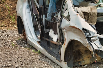 A heavily damaged and rusted white car is positioned at the side of a gravel path. The doors are missing and the interior is exposed, with some debris and cables visible. The metallic parts show significant corrosion.