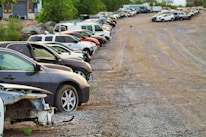 A row of parked cars on a gravel lot with trees in the background. Some cars appear to be damaged or missing parts, and the area has an industrial feel with a warehouse visible at the edge.